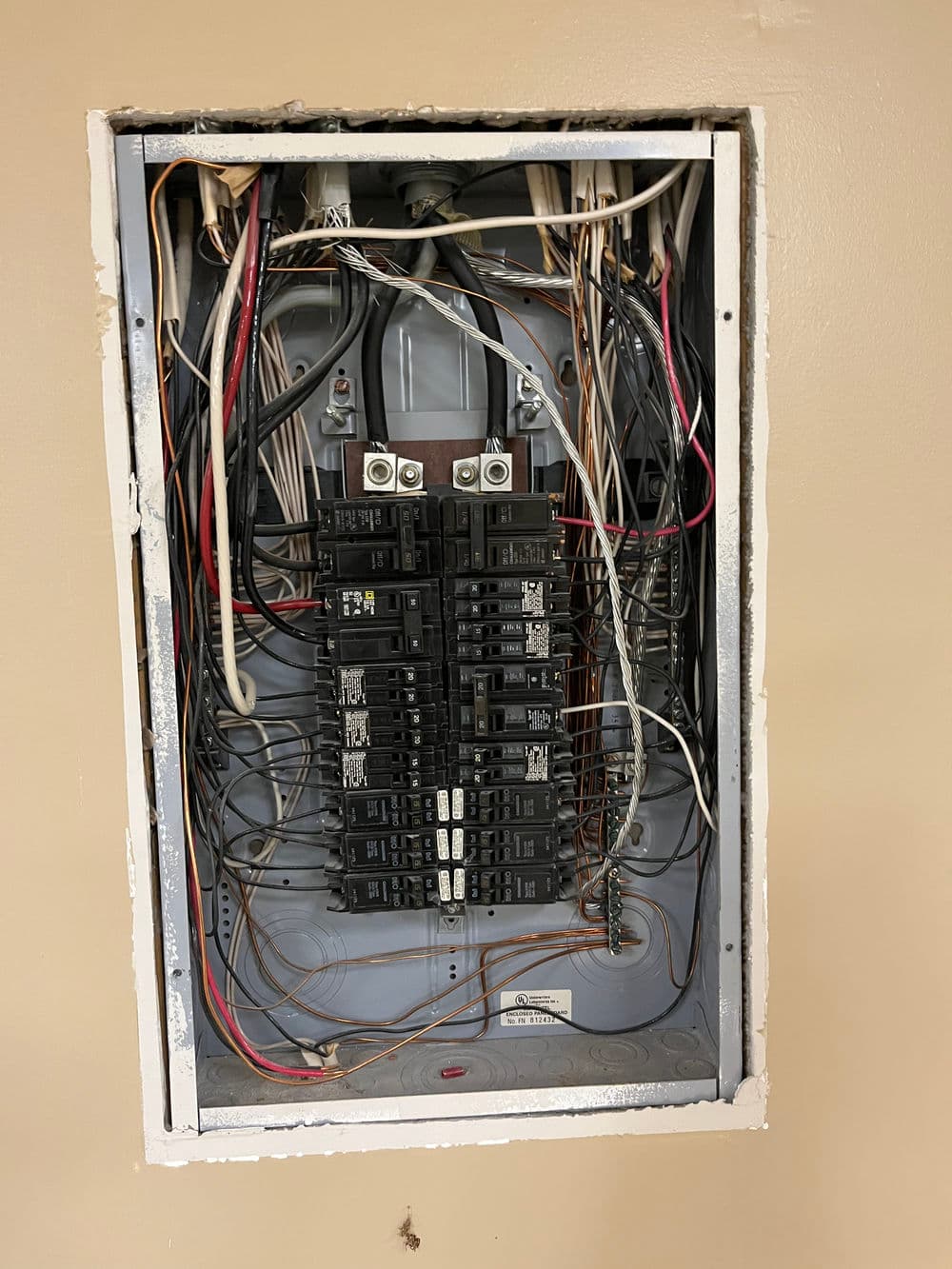 Electrical panel with circuit breakers, exposed wires, and uneven wall opening.